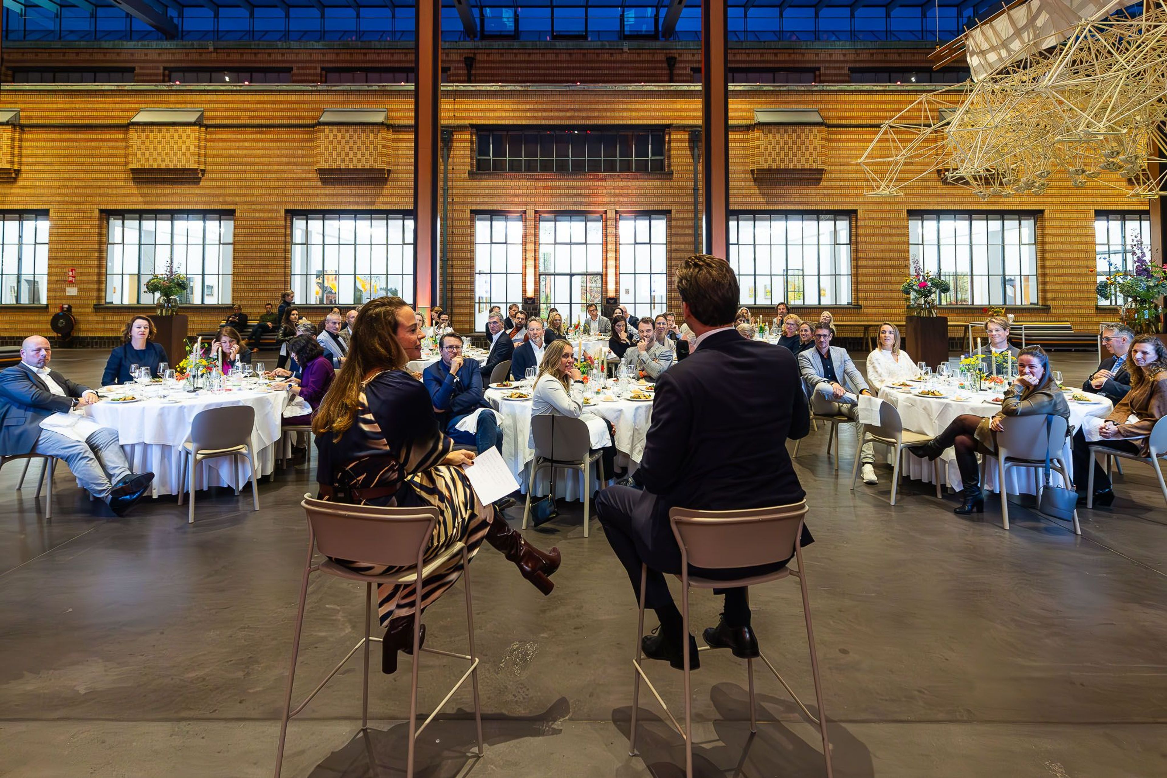 Zakelijke bijeenkomst met sprekers en publiek aan tafels.