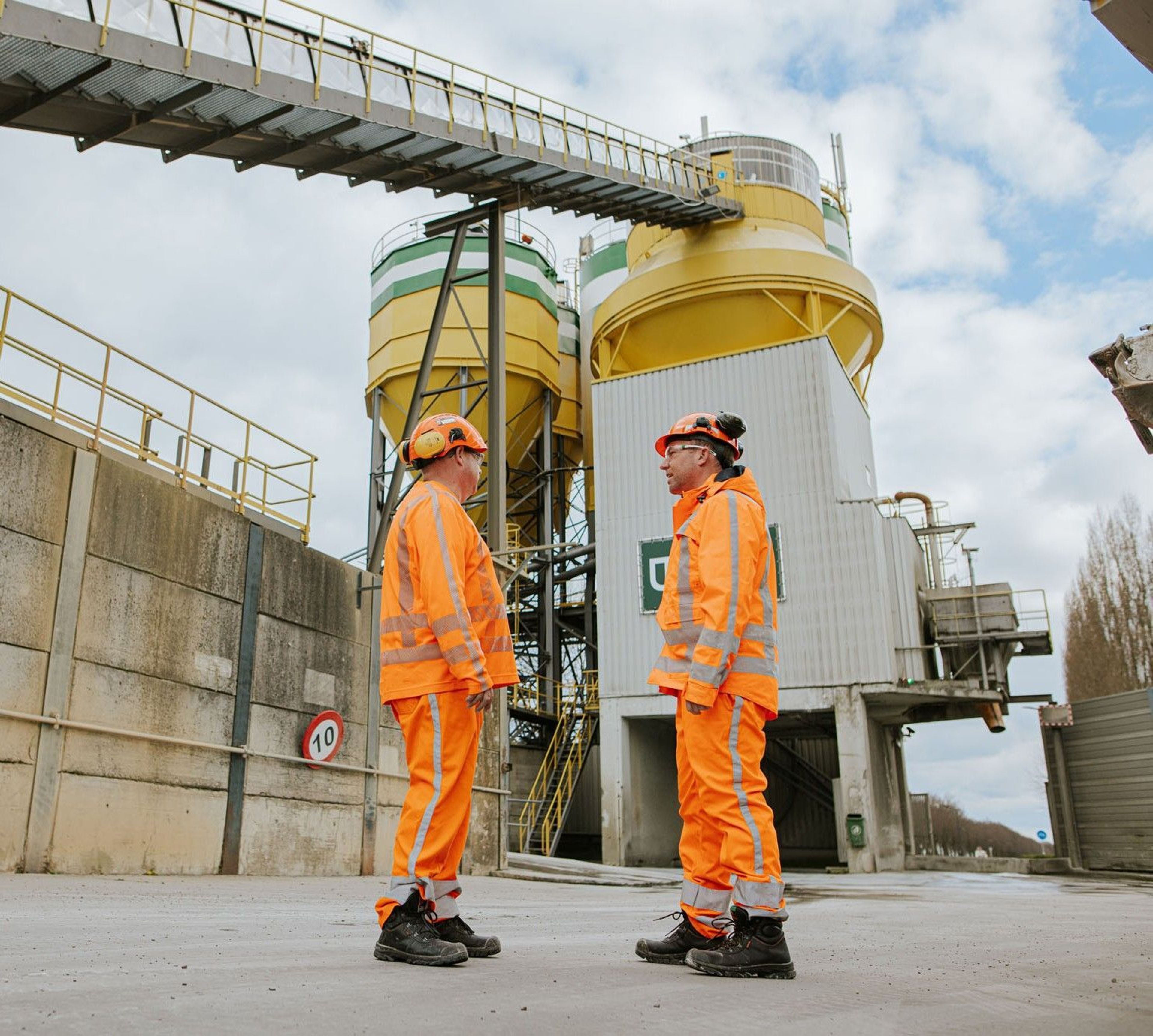 Twee werklieden in oranje veiligheidskleding bij een fabriek.