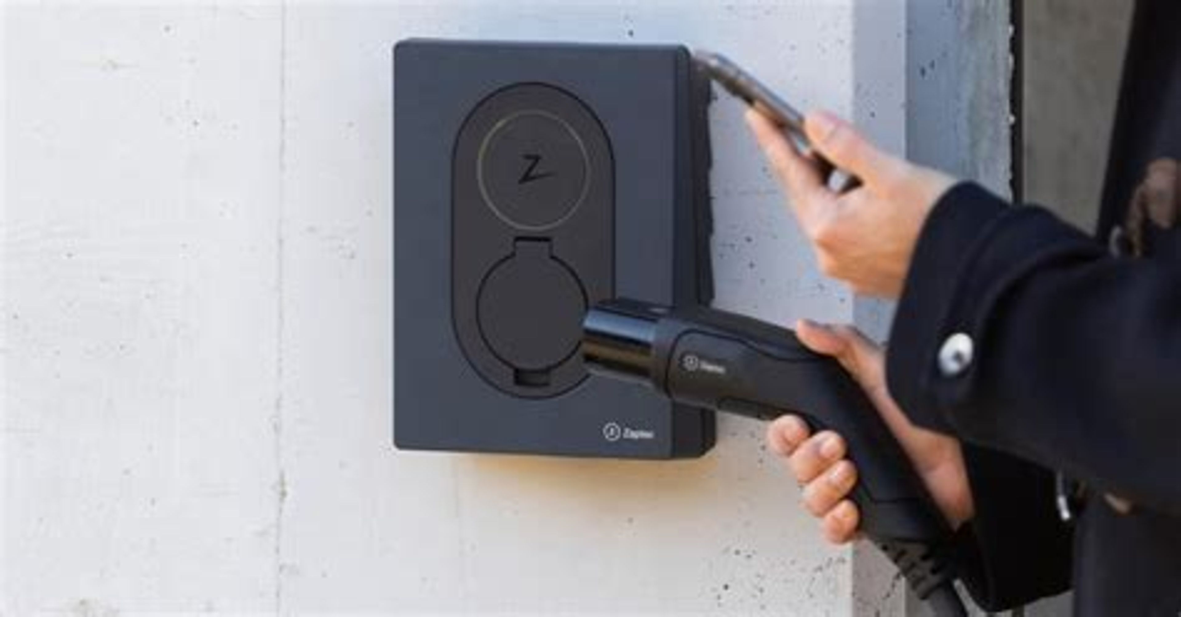 Hand die een elektrische auto oplaadkabel aansluit op een laadpaal wandmodel bij een gebouw.
