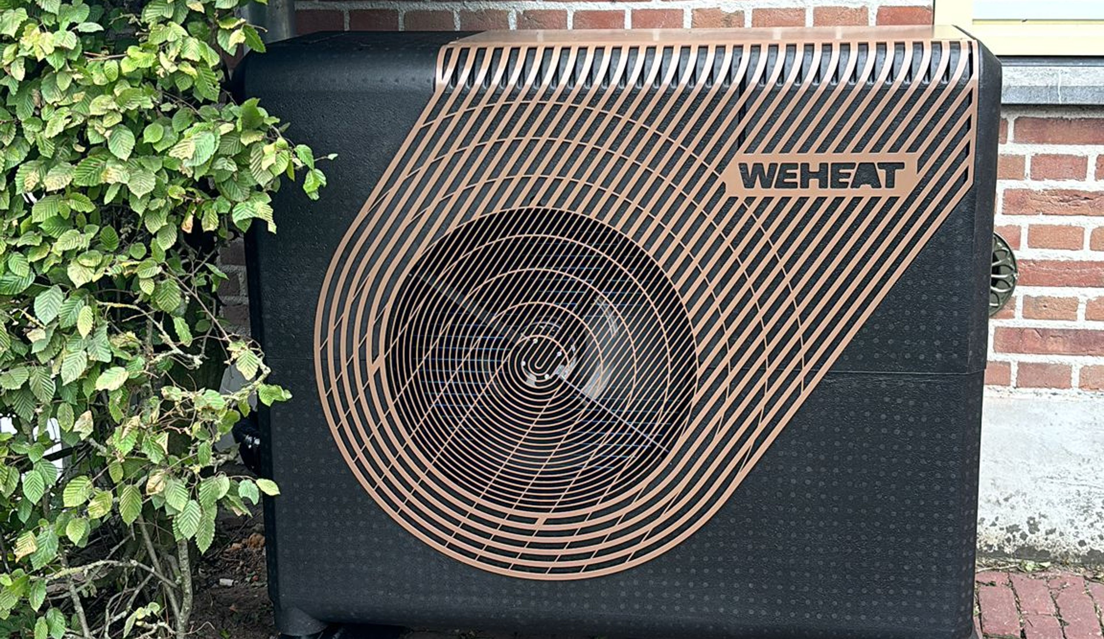 Een zwart weheat warmtepomp apparaat geplaatst naast een gevel met rode bakstenen en groene klimop.