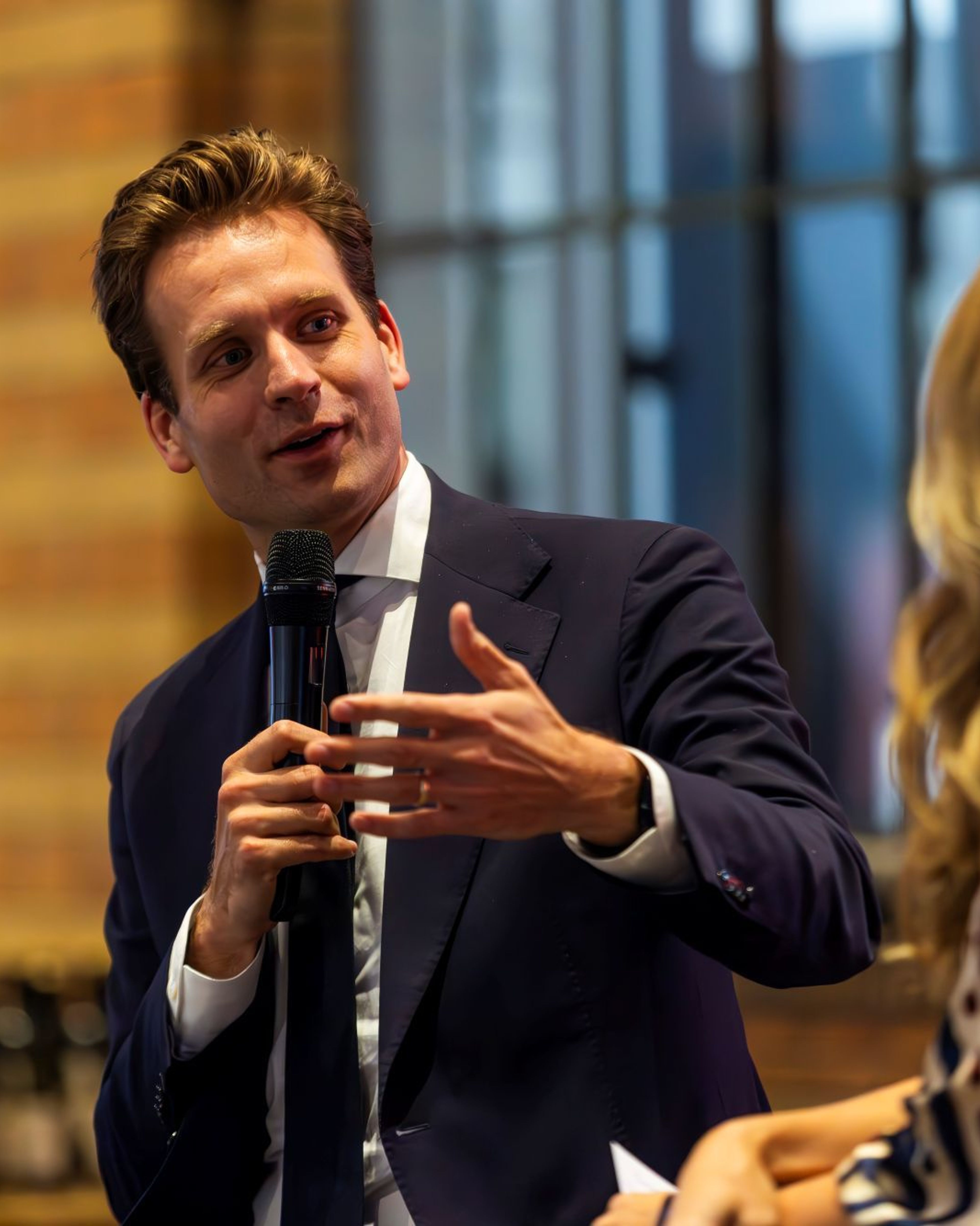 Man met microfoon in gesprek, vrouw op de achtergrond.