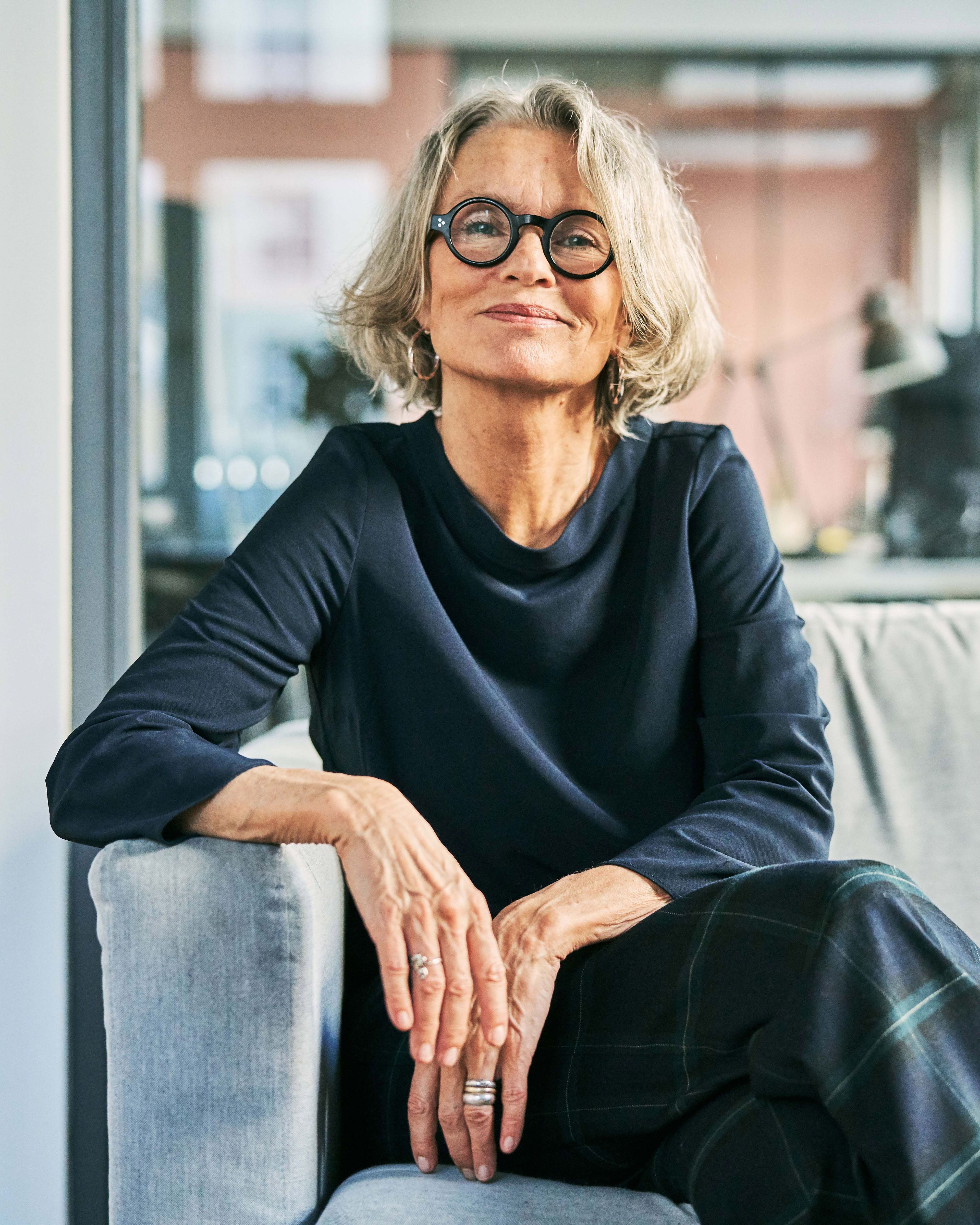 Een vrouw met grijs haar en ronde bril, zittend op een bank.