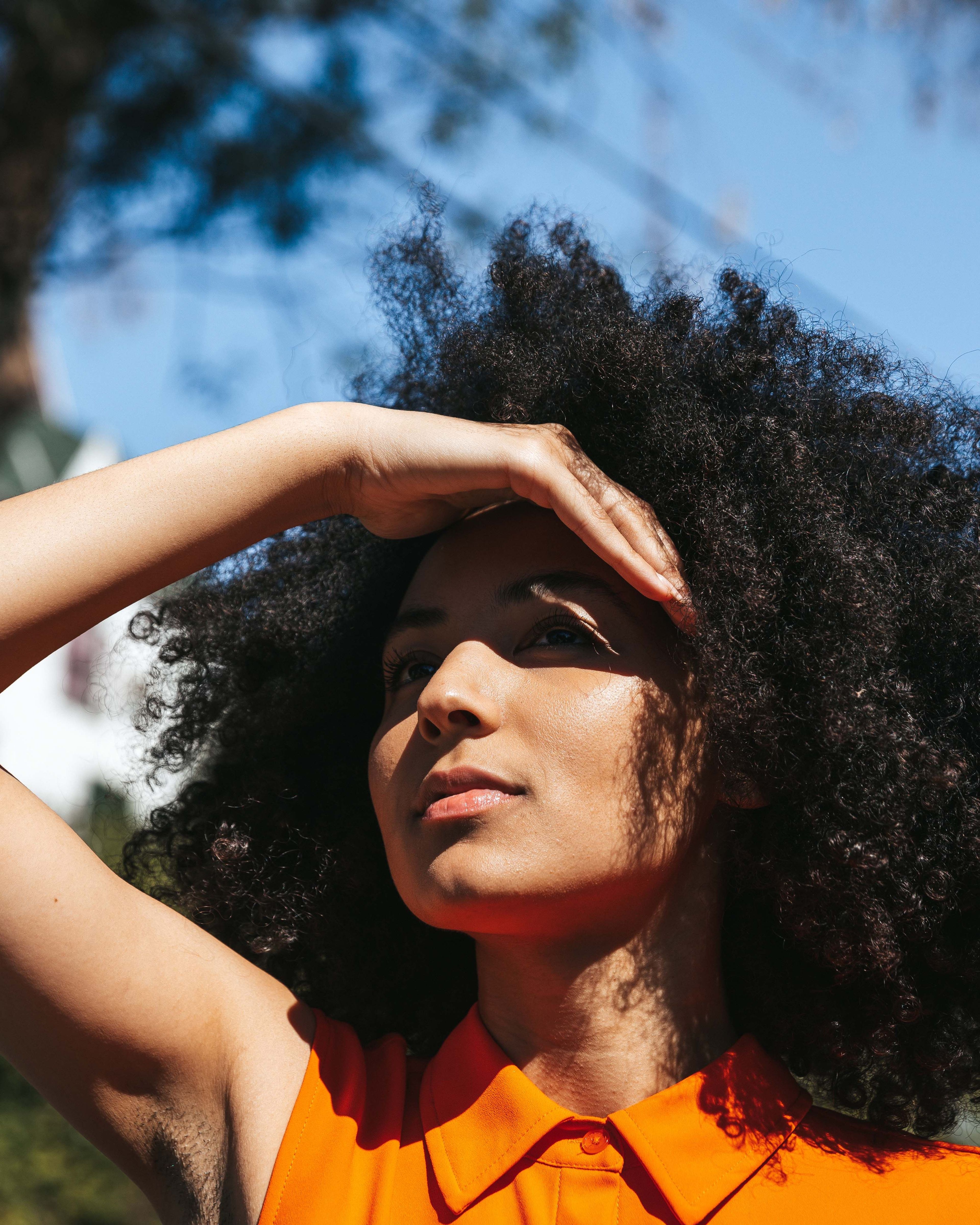 Vrouw met krullend haar in oranje blouse, onder een zonnige lucht.