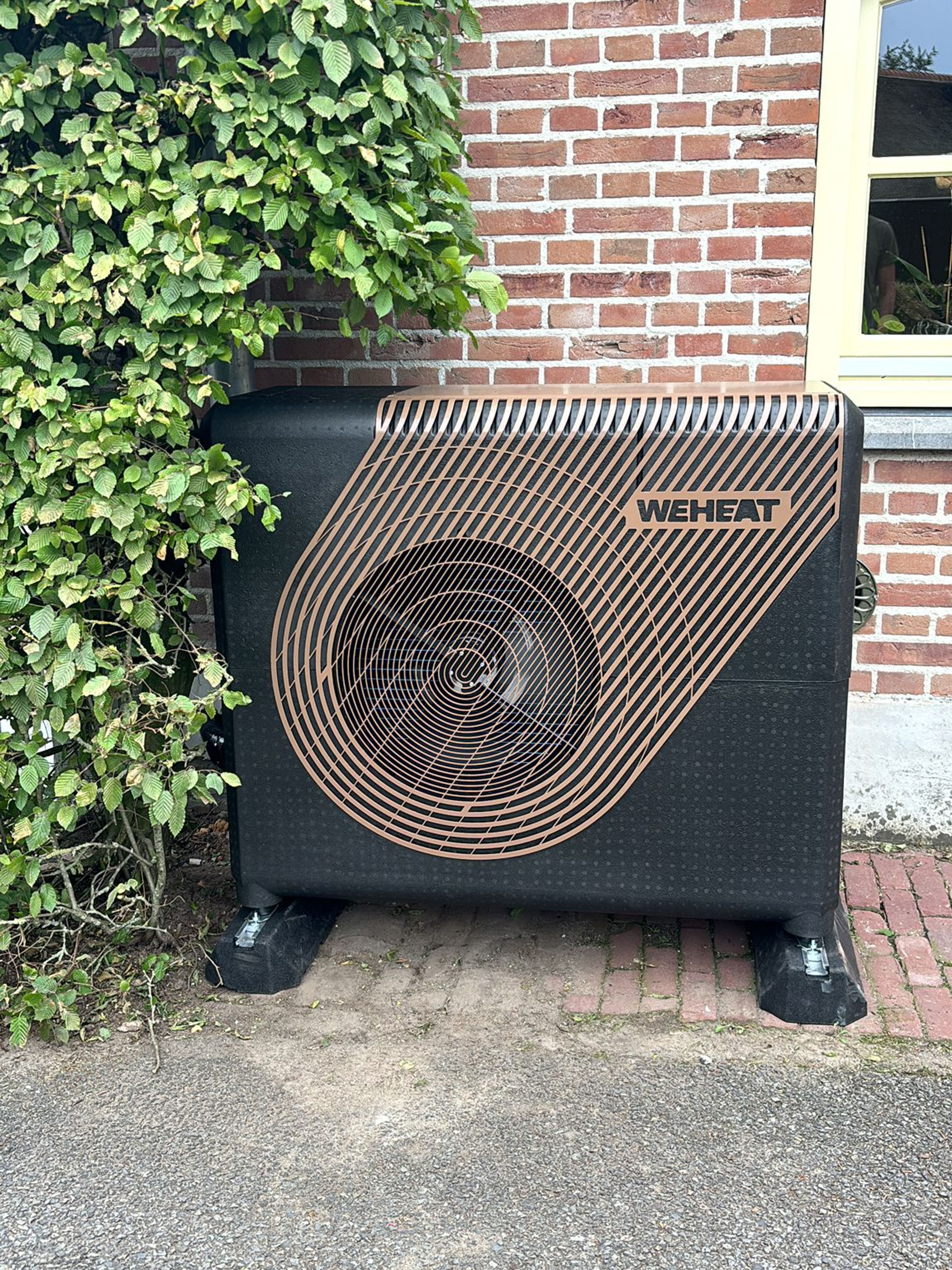 Een zwart weheat warmtepomp apparaat geplaatst naast een gevel met rode bakstenen en groene klimop.