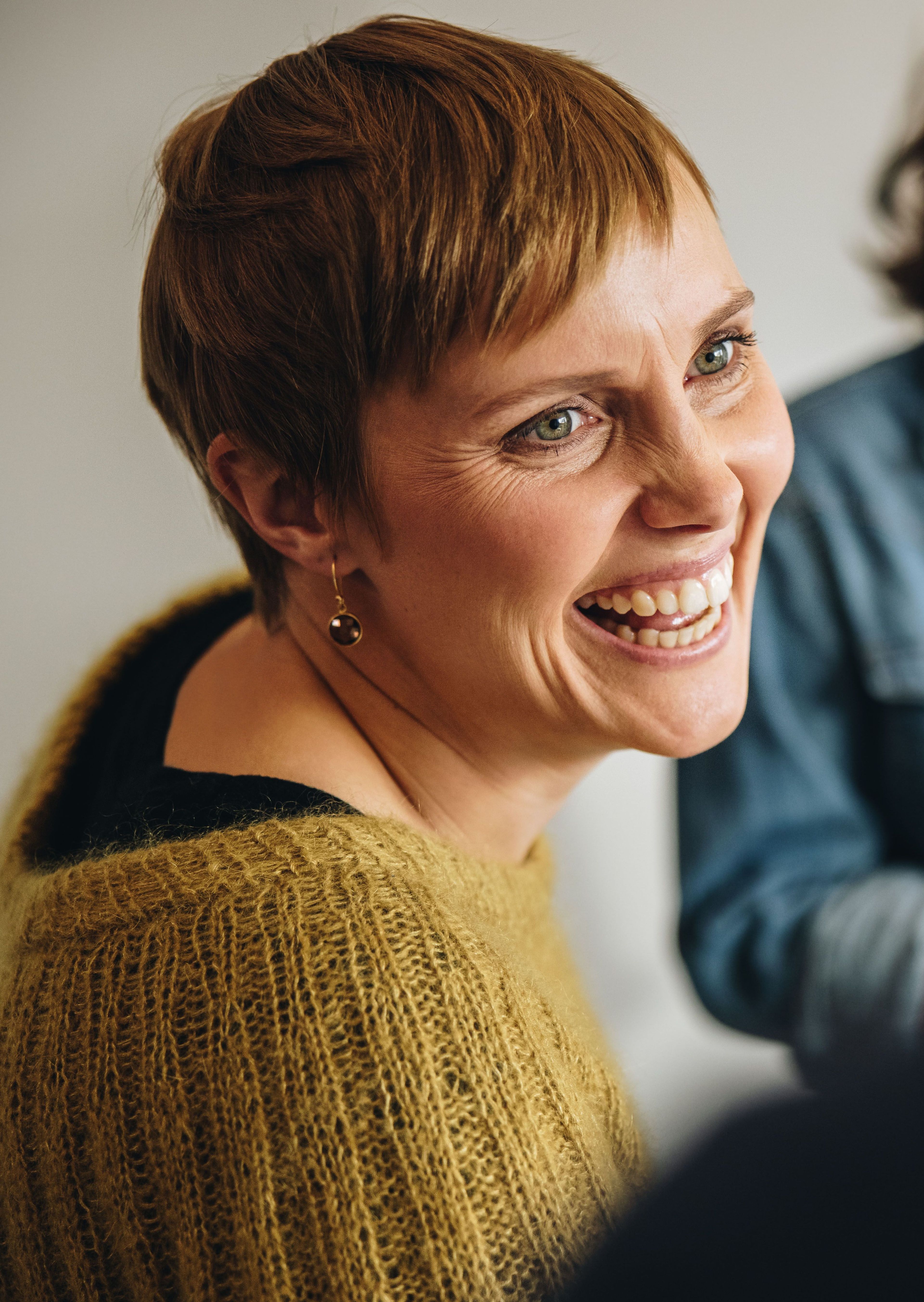Vrouw met kort haar en een glimlach, gekleed in een gouden trui.